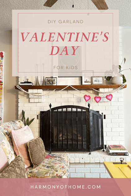 DIY Valentine garland craft for kids featuring 3 red hearts hanging from a wooden fireplace mantle with white brick. 