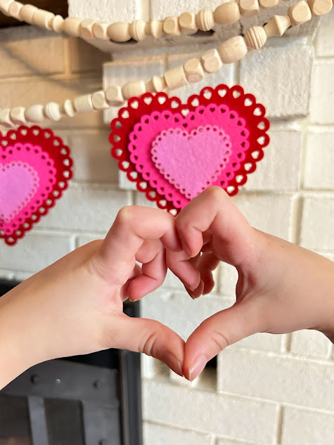 A child's hands make a heart shape in front of a beaded garland craft with red and pink felt hearts in the background.