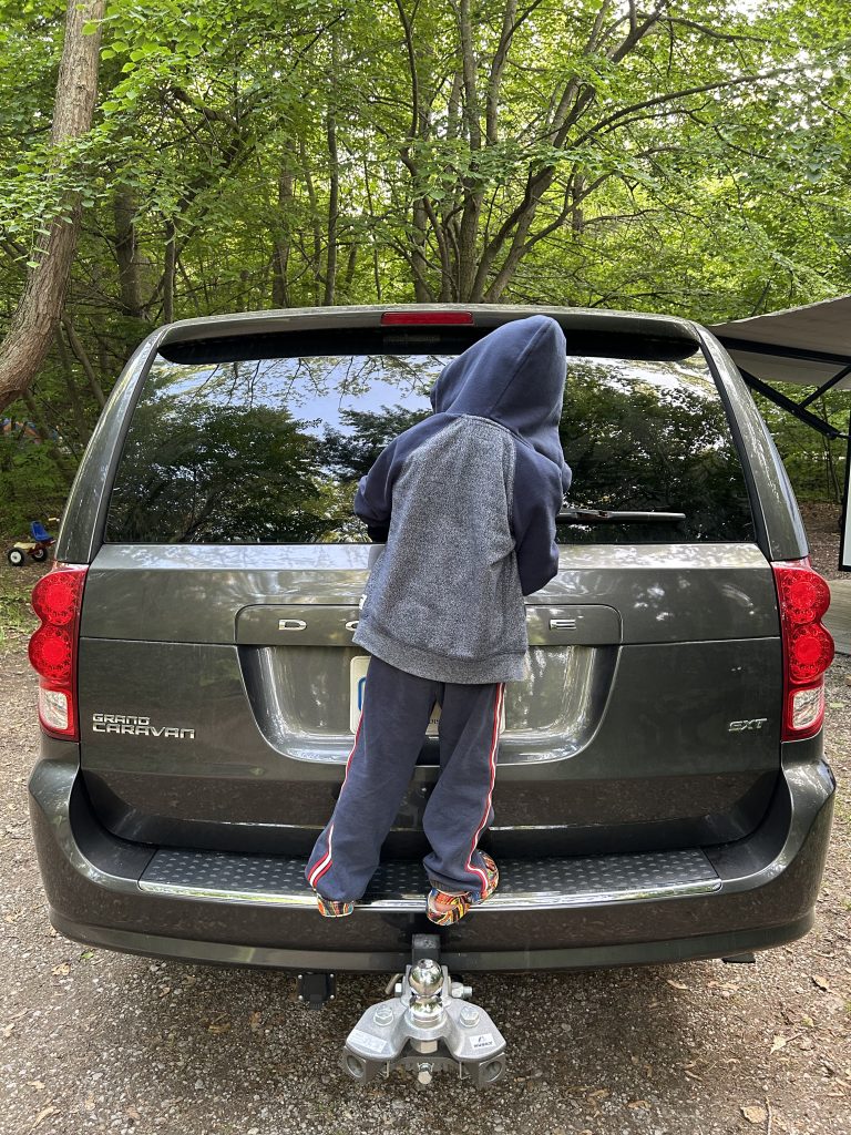 Child standing on the back bumper of a van looking through the back window parked in a forest setting